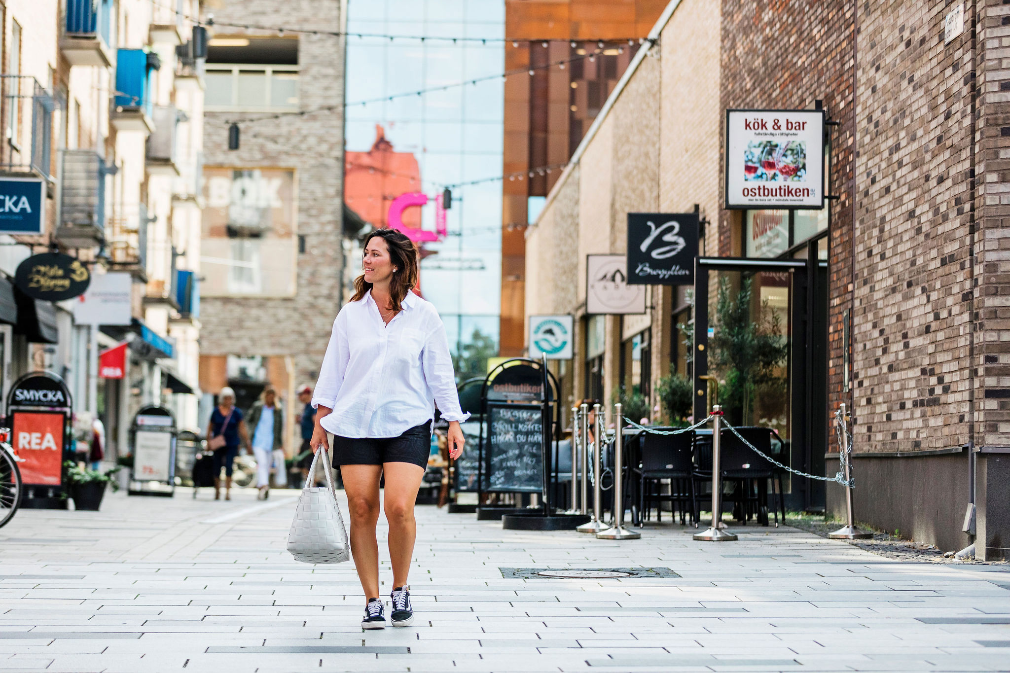 Mörkhårig ung kvinna iklädd vit skjorta, svarta shorts och sneakers bär på en vit väska på promenad i Mölndals innerstad. En gata med butiker på sidorna. 