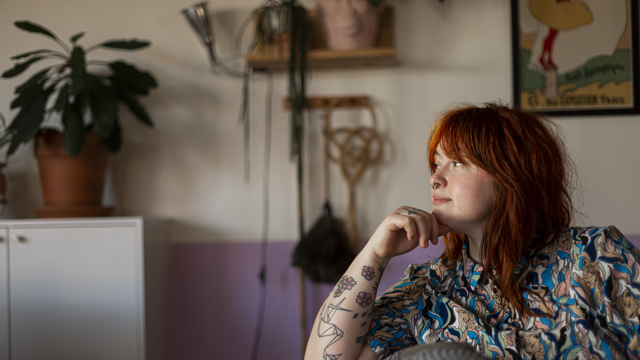 Närbild på ung rödhårig kvinna funderar sittandes i sin soffa, i bakgrunden syns diverse städredskap hängandes på väggen. 