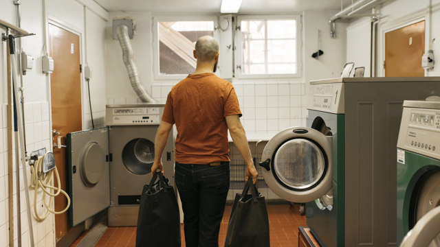 Tunnhårig man bakifrån bärandes två kassar med tvätt i en tvättstuga. 