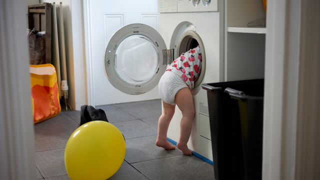 Ett badrum med en tvättmaskin i fokus där ett litet blöjbarn kikar in med hela överkroppen i tvättmaskinenslucka. Några ballonger ligger på golvet bredvid. 