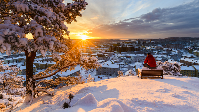 En person sitter på ett högt berg omgiven av snö och blickar ut över Mölndal i solnedgången.