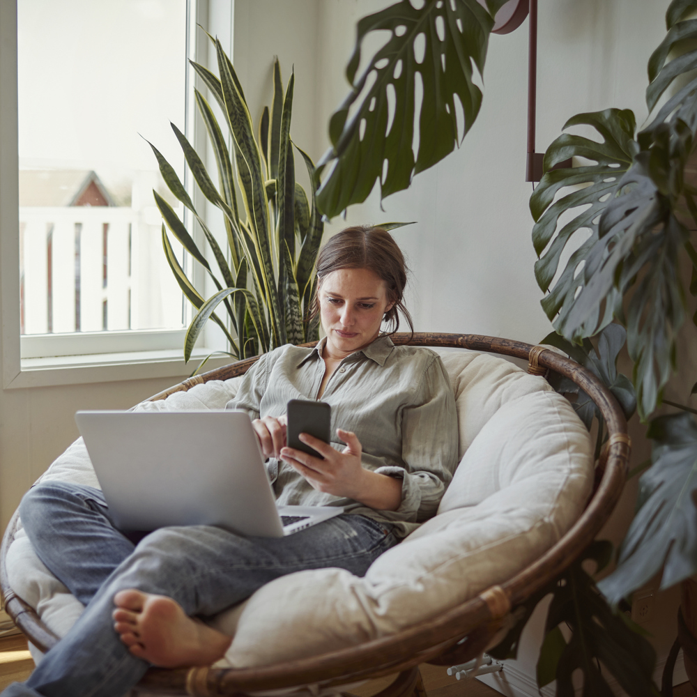 En ung kvinna sitter i en rottingfotölj med en laptop i knät och en smartphone i handen i sitt vardagsrum. 