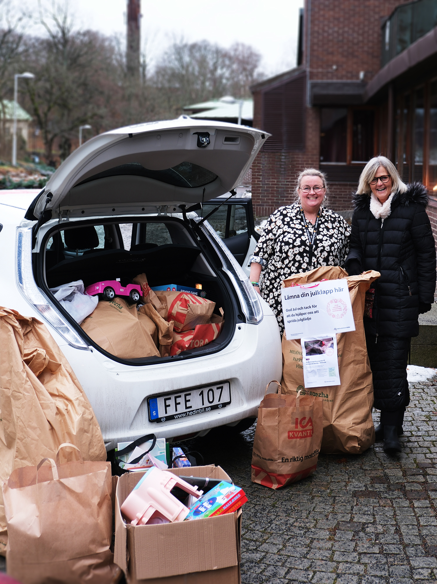 två kvinnor står bredvid en bil med öppet bagage fylld med julklappar för insamlingen Julgåvan utanför församlingshemmet i Mölndal 