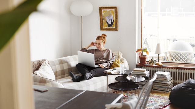Ung kvinna sitter i sin soffa med en dator i knät i ett ljust vardagsrum. 